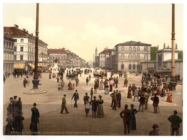 Schwarz-weiß-Foto einer belebten Münchner Straße in den frühen 1900er Jahren mit Fußgängern, einem Pferdewagen, Gebäuden, Bäumen und einem sichtbaren Himmel, mit Text am unteren Rand.