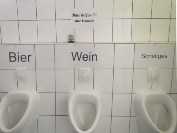 Drei weiße Toiletten an einer Wand montiert mit Schildern darüber, die "Bier Wein" und "Sonstiges" lesen.