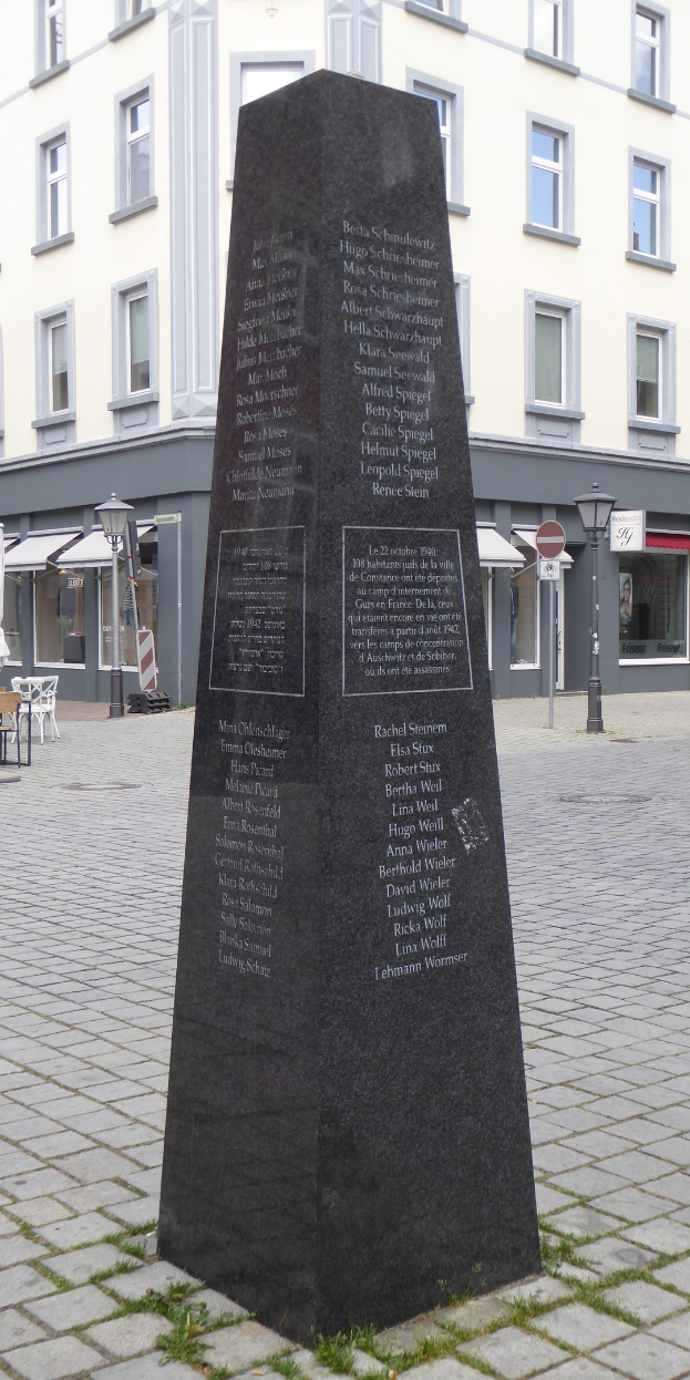 Ein Denkmal mit Inschrift steht in einem Stadtplatz umgeben von Gebäuden, Straßenmöblierung und Menschen unter einem sichtbaren Himmel.