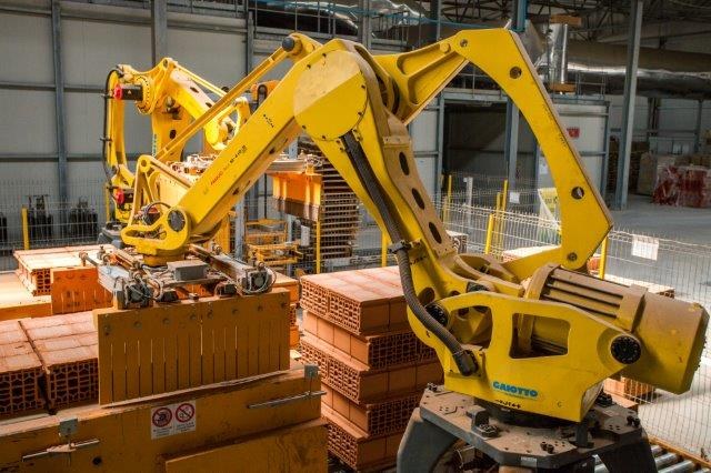 Großes Industriegebäude mit gestapelten Kisten, Kränen an der Decke, Zaun, Versorgungsmasten und einer fernen Wand in einer Fertigungseinrichtung.