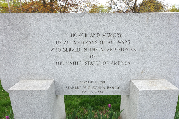 Gedenkstein mit einer Inschrift für US-Streitkräfte-Veteranen umgeben von Pflanzen, Blumen, Gras, Bäumen und einem bewölkten Himmel.