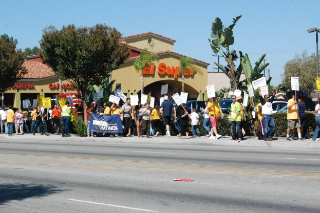 Eine Gruppe von Menschen mit Schildern und Transparenten, die an einem Gebäude mit Namensschildern, kleinen Büschen und Autos vorbeigehen.
