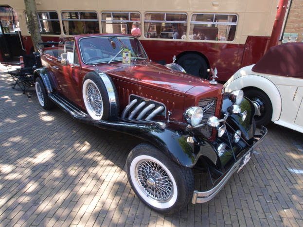 Ein Vintage-Auto neben einem Doppeldeckerbus auf einer Kopfsteinpflasterstraße, mit Stühlen und einem Baum im Vordergrund, einem Gebäude mit Fenstern im Hintergrund und Menschen im Bus zu sehen.