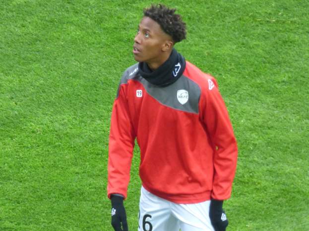 Ein junger Mann in einer roten Jacke und weißen Shorts steht auf einem Fußballfeld, trägt Baseballhandschuhe und sieht entschlossen nach rechts.