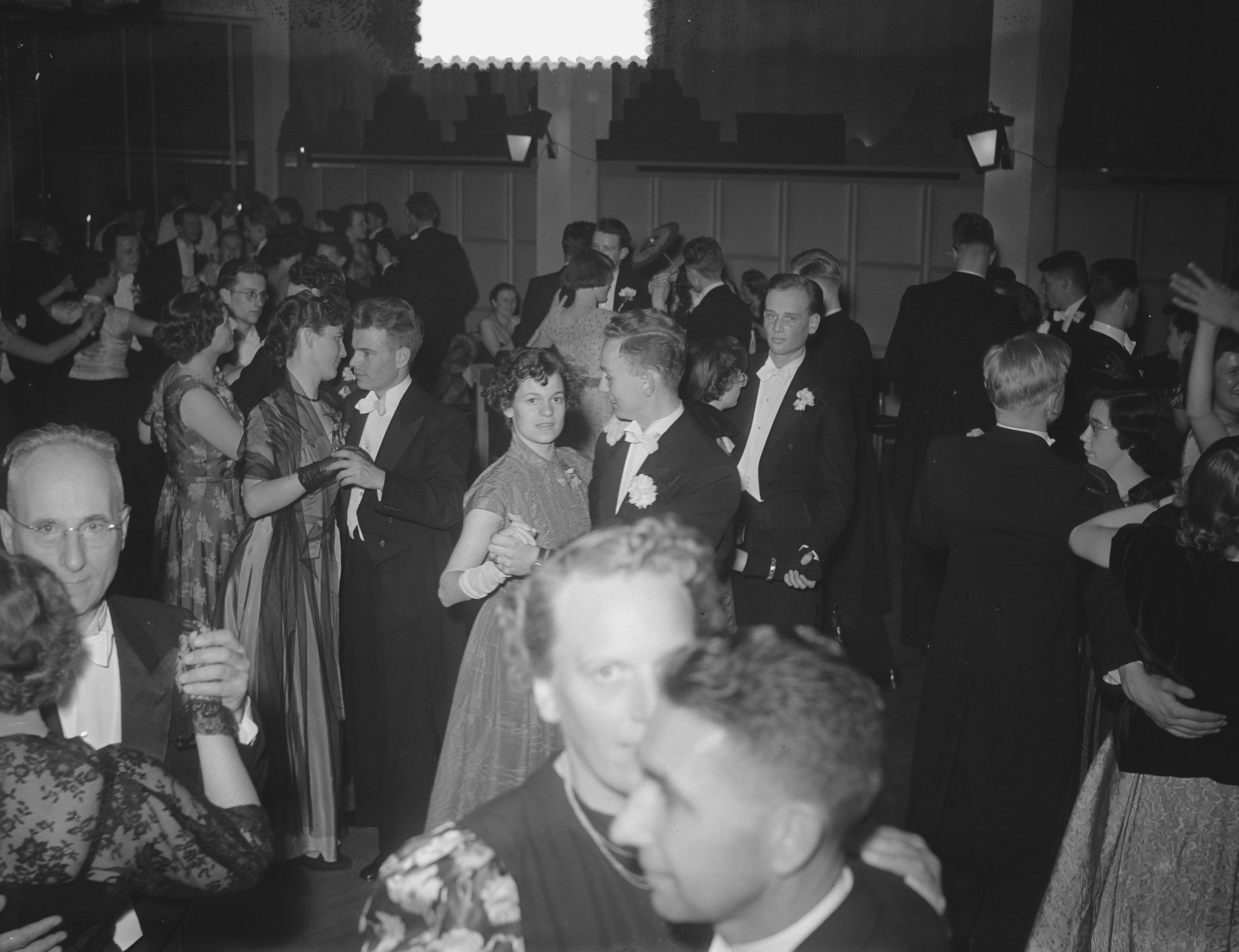 Eine Gruppe von Menschen verschiedenen Alters, die auf einer Party tanzen, einige halten Gl├Ąser, mit beleuchteter Wand im Hintergrund.