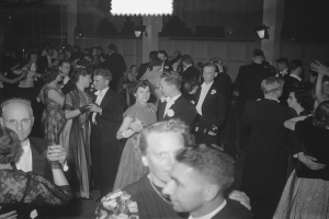 Eine Gruppe von Menschen verschiedenen Alters, die auf einer Party tanzen, einige halten Gl├Ąser, mit beleuchteter Wand im Hintergrund.