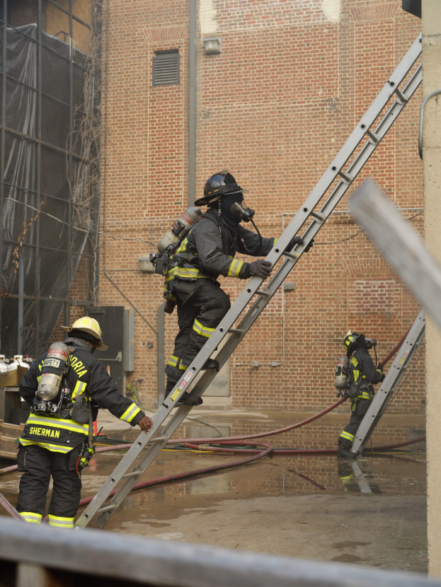Feuerwehrleute mit Helmen und Lufttanks klettern an einer Leiter hoch vor einem Backsteingebäude mit Rohren auf dem Boden und einer Metallstange unten, mit einem weiteren Gebäude mit Fenstern und einem Netz im Hintergrund.