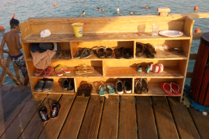 Eine hölzerne Bank auf einem Dock mit verschiedenen Schuhen, einem Becher, einer Flasche, einem Teller, einem Glas und anderen Gegenständen, daneben ein Müllcontainer und schwimmende Menschen im Hintergrund.
