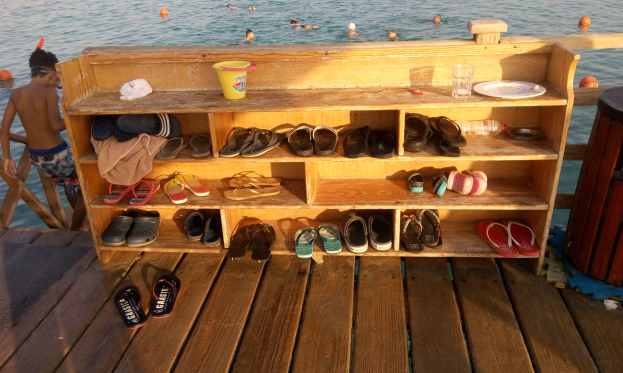 Eine hölzerne Bank auf einem Dock mit verschiedenen Schuhen, einem Becher, einer Flasche, einem Teller, einem Glas und anderen Gegenständen, daneben ein Müllcontainer und schwimmende Menschen im Hintergrund.