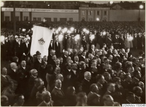 Schwarzes und weißes Foto einer großen Menge vor einem Gebäude, mit einer Person in der Mitte, die eine Fahne hält, sowie Polen und Lichter im Hintergrund und Text unten, der "Gustav Klimts Rede an der Berliner Mauer" lautet.