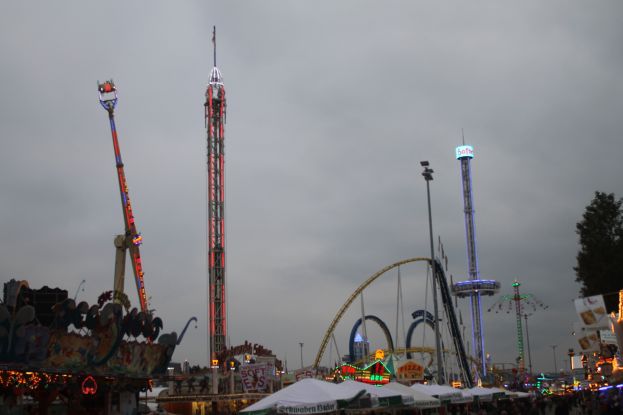 Eine Menschenmenge versammelt sich um einen hell erleuchteten Karneval in der Nacht, mit beleuchteten Fahrgeschäften, Pfählen und Bäumen unter einem bewölkten Himmel.