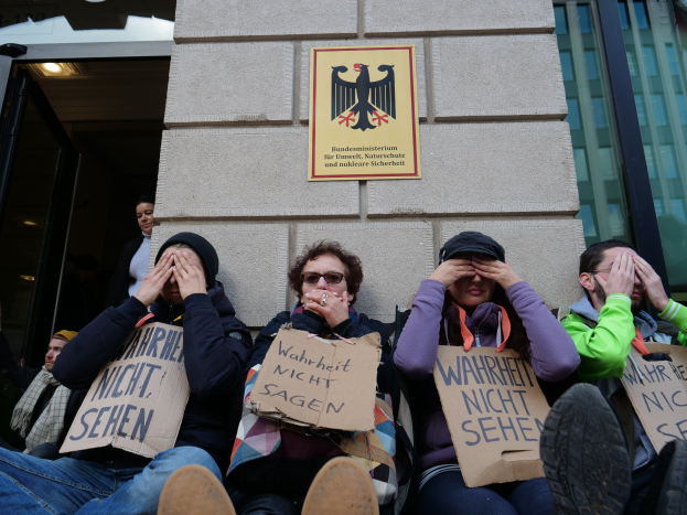 Eine Gruppe von Menschen sitzt vor einem Gebäude und hält Schilder hoch, während einige ihre Gesichter mit den Händen bedecken.
