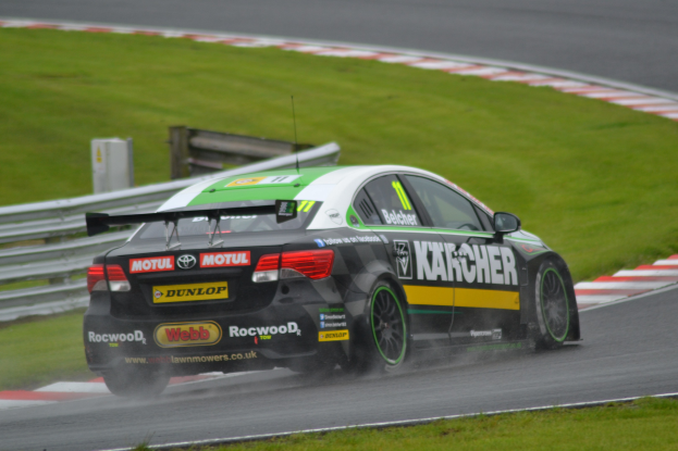 Ein Auto mit Text auf der Seite fährt auf einer regennassen Rennstrecke, die von Gras umgeben ist, mit einem Geländer und einer hölzernen Konstruktion im Hintergrund.