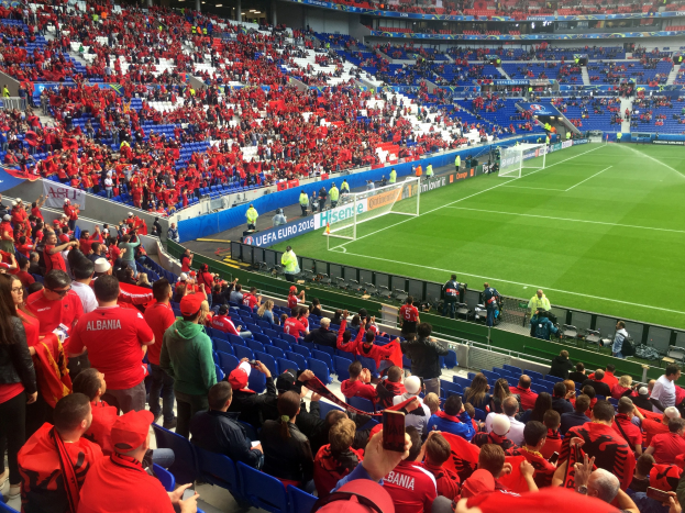 Ein volles Stadion mit Zuschauern, einige sitzen und einige stehen, die ein Fußballspiel beobachten, mit sichtbaren Torpfosten, Bannern und Deckenlampen unter einer Dachkonstruktion.