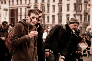 Eine Gruppe von Menschen in Zombie-Kostümen geht eine Straße entlang, einige tragen Kostüme und einer hält eine Kamera, im Hintergrund ist ein Gebäude zu sehen.